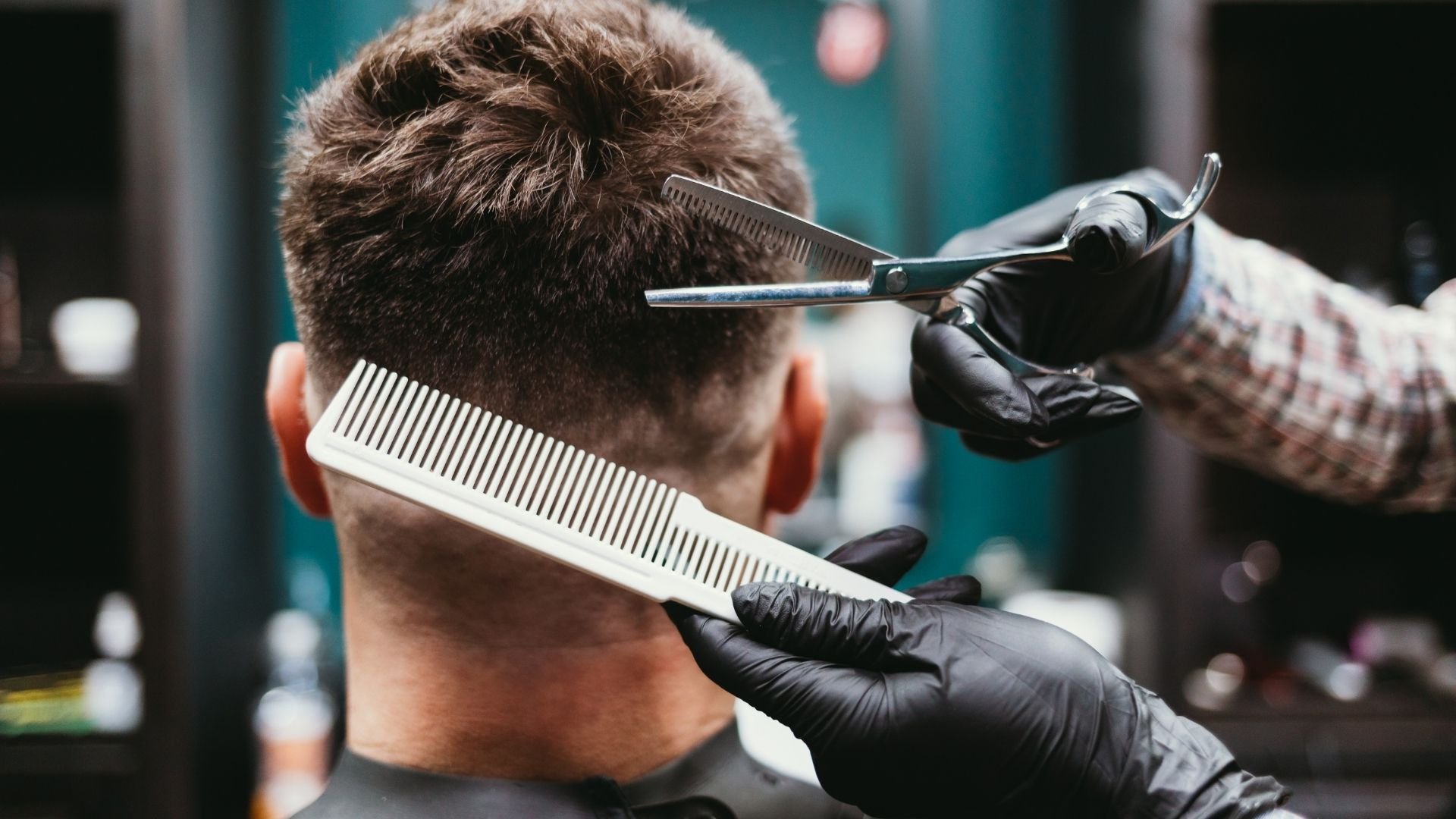 Barber wearing black gloves cutting client's hair with scissors and comb.