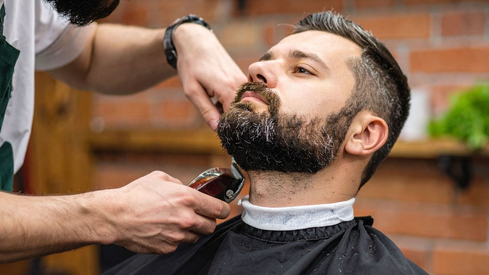 Barber trimming bearded man's beard with clippers in professional salon setting